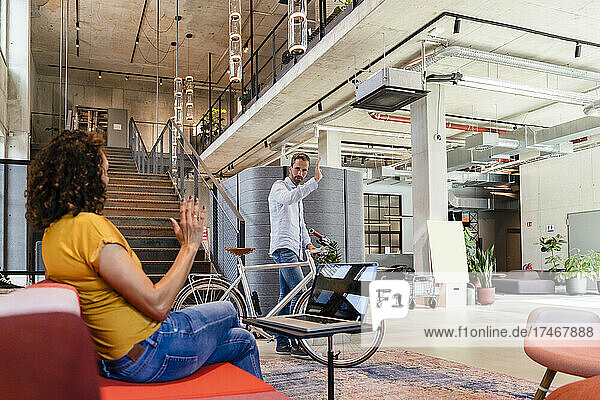 Businessman with bicycle waving hand to female coworker in office
