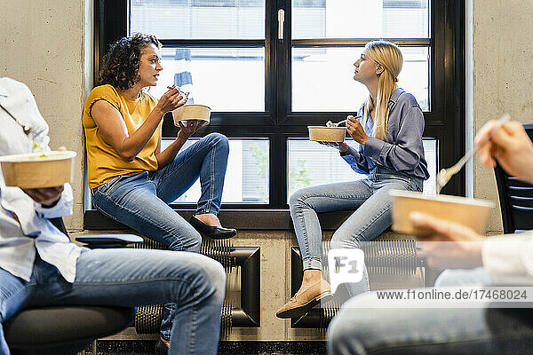 Businesswomen having food at window sill with colleagues in office