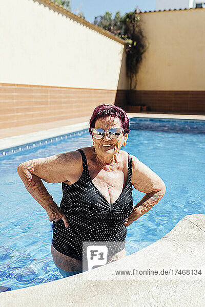 Smiling senior woman with hand on hip standing in swimming pool during vacation