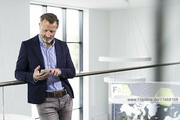 Businessman using mobile phone while leaning on railing in office