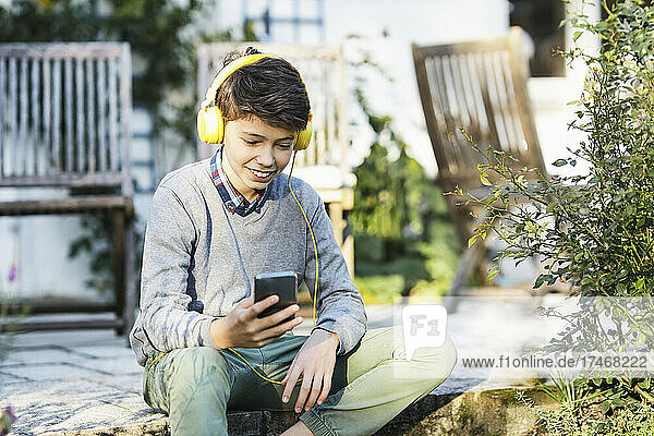 Smiling boy wearing headphones using smart phone at backyard
