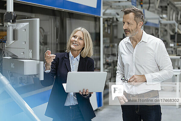 Smiling female professional gesturing while discussing with male colleague over machinery in factory