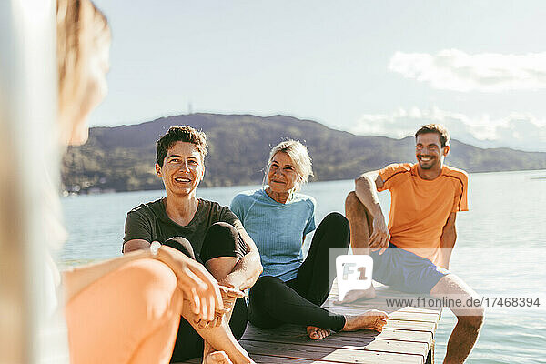 Männliche und weibliche Freunde reden miteinander  während sie auf dem Steg am See sitzen