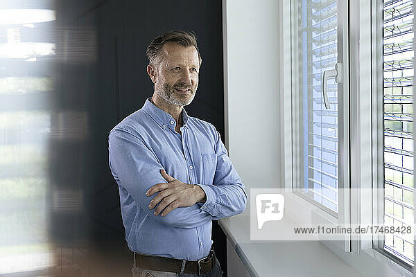 Businessman looking through window while standing with arms crossed in office