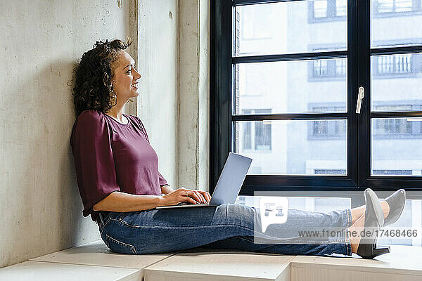 Smiling businesswoman with laptop looking through window in office