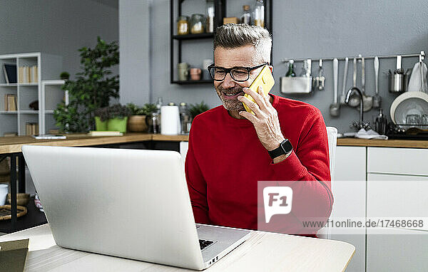 Mature man wearing eyeglasses talking on smart phone while sitting with laptop at table
