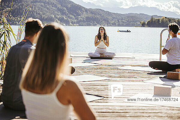 Female instructor with hands clasped conducting yoga class on jetty by lake