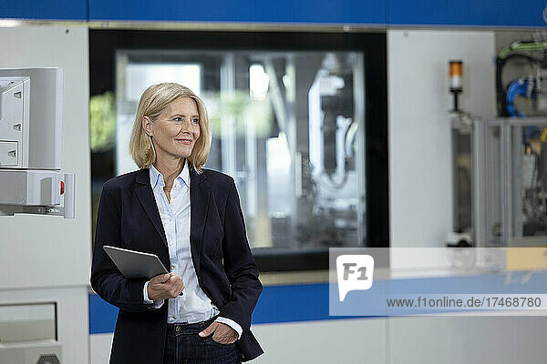 Mature businesswoman with hand in pocket holding digital tablet at factory