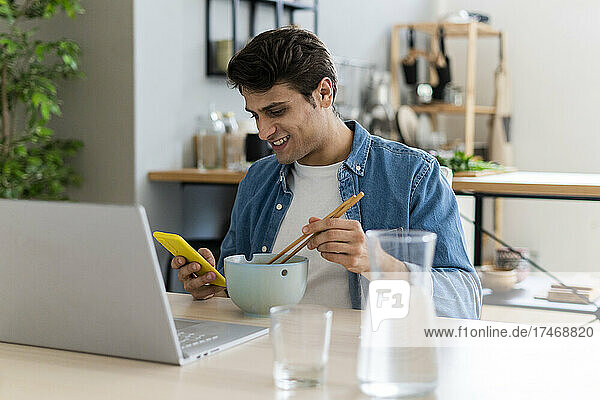 Smiling man using mobile phone while having food at home