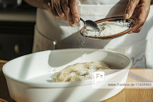 Woman putting grated coconut on dough in bowl at home
