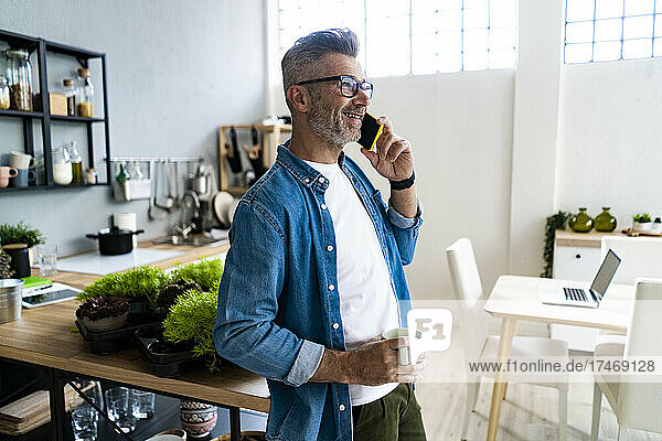 Mature man holding coffee cup while talking on smart phone at home