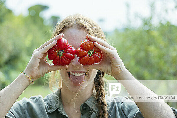 Playful female farmer holding tomatoes in front of eyes and sticking out tongue