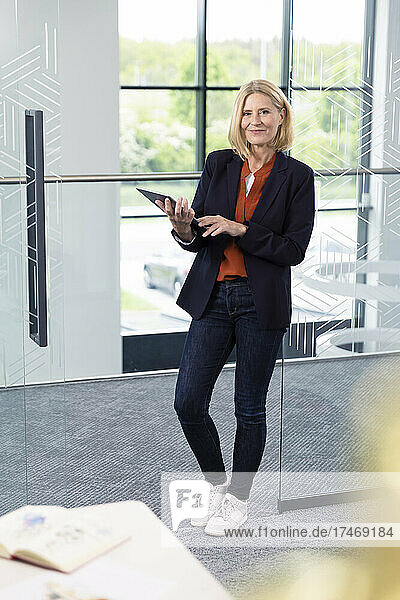 Mature businesswoman with digital tablet leaning on glass door