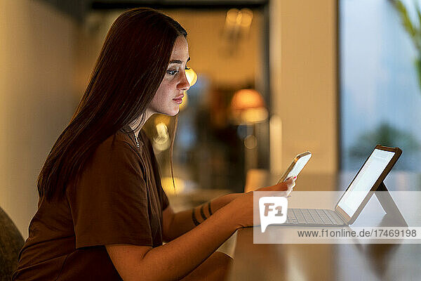 Young woman using smart phone by laptop at table