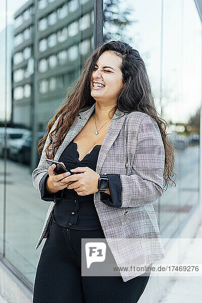 Cheerful businesswoman with smart phone laughing in front of glass