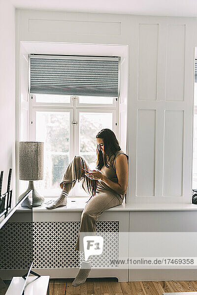 Woman using smart phone sitting on window sill at home