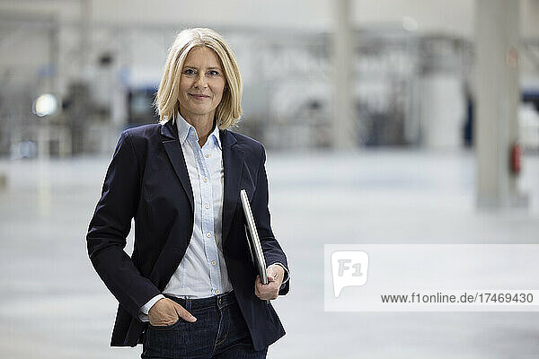 Smiling mature businesswoman standing with hand in pocket while holding laptop in industry
