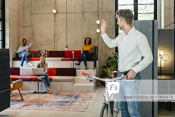 Businessman with bicycle waving hand to colleagues in office