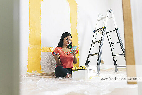 Smiling young woman with paint roller using smart phone in living room