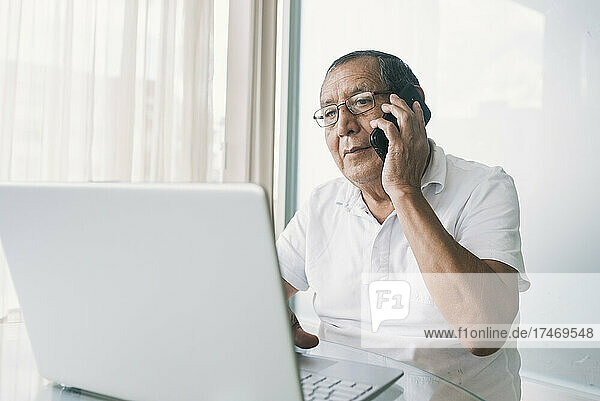 Senior businessman with laptop talking on mobile phone at home office