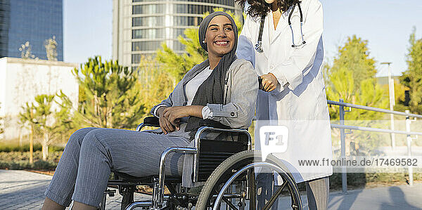 Ärztin schiebt Rollstuhl einer lächelnden behinderten Frau auf Fußweg