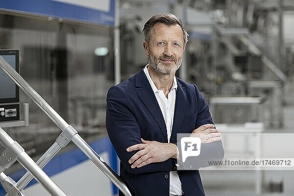 Mature businessman standing with arms crossed by railing in factory