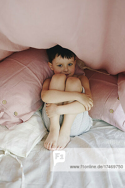 Boy hugging knees below blanket at home