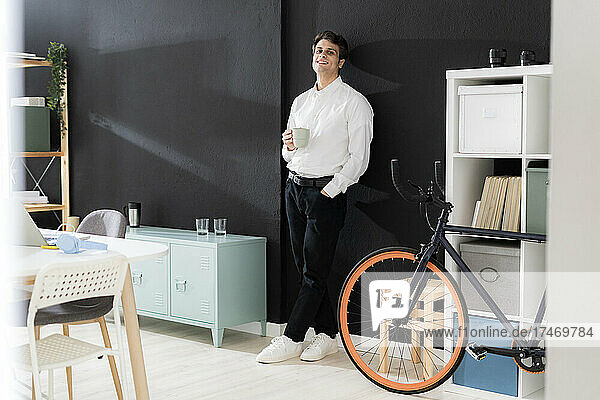 Young businessman with coffee cup leaning on black wall in creative office
