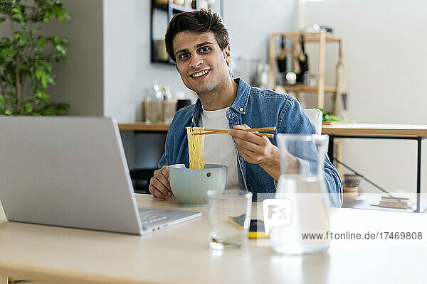Young man sitting with laptop eating noodles at table