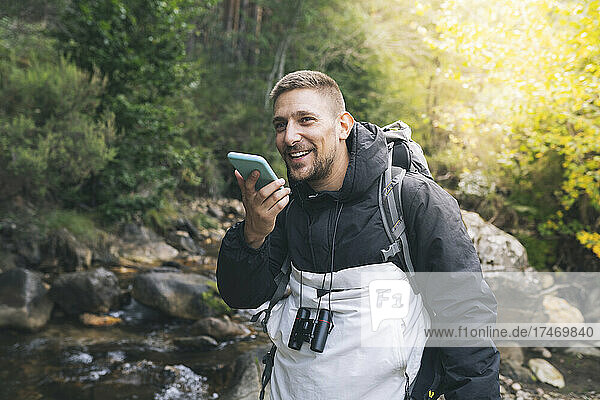 Smiling male tourist talking on smart phone through speaker at forest