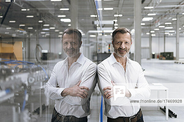 Smiling businessman standing with arms crossed while leaning on glass in factory
