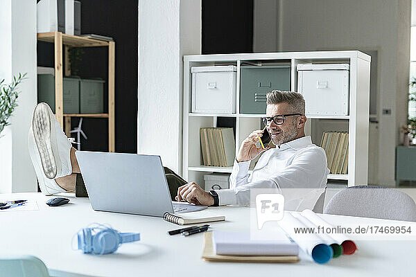 Geschäftsmann spricht am Smartphone  während er im Büro einen Laptop benutzt