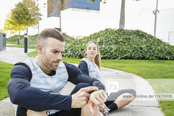 Sportsman checking wristwatch while sitting by female friend at park