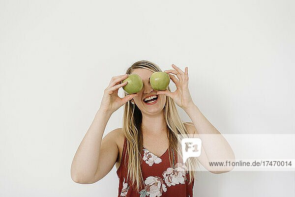 Cheerful blond female nutritionist covering eyes with apples in front of white wall