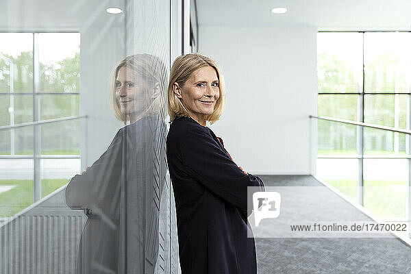 Mature businesswoman with arms crossed leaning on glass in corridor