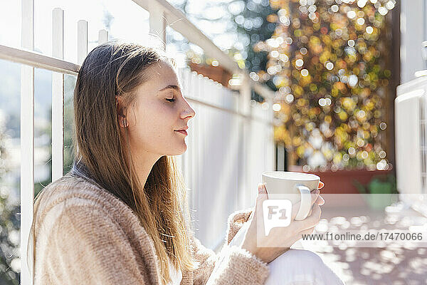 Junge Frau hält Kaffeetasse in der Hand  während sie mit geschlossenen Augen auf dem Balkon sitzt