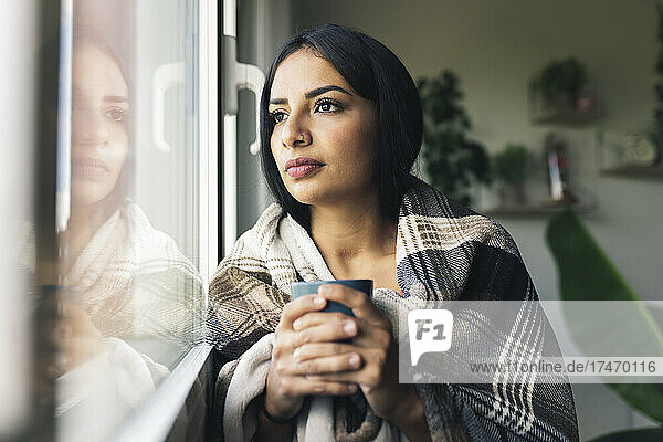 Young woman with coffee mug looking through window at home