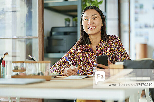 Lächelnde Geschäftsfrau mit Smartphone sitzt am Arbeitsplatz