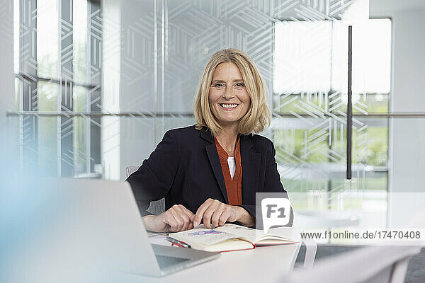 Lächelnde blonde Geschäftsfrau sitzt mit Tagebuch am Schreibtisch