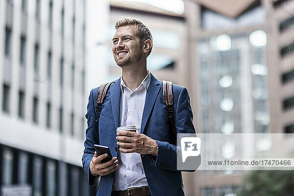 Happy young male professional with mobile phone and disposable cup in city