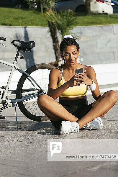 Junge Sportlerin benutzt Smartphone  während sie mit dem Fahrrad auf dem Fußweg sitzt