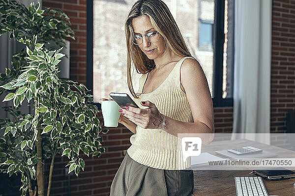Businesswoman using mobile phone while holding mug at desk