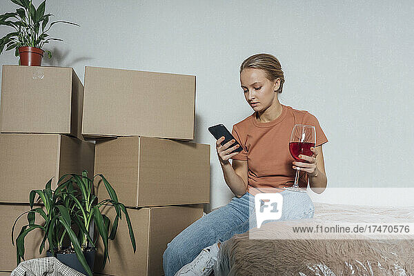 Woman with wineglass using mobile phone in relocated apartment