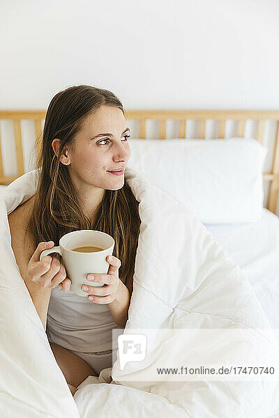Young woman holding coffee cup while wrapped in white blanket at home