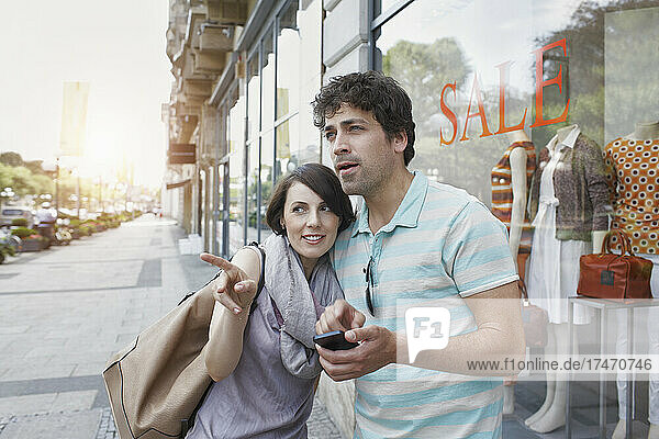 Frau zeigt  während Freund mit Smartphone neben Geschäft auf Fußweg steht
