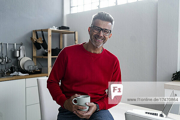 Lächelnder Mann mit Kaffeetasse sitzt zu Hause