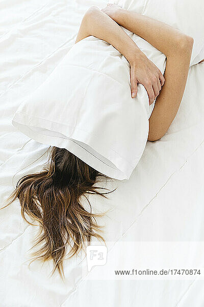 Young woman covering face with white pillow while lying on bed at home