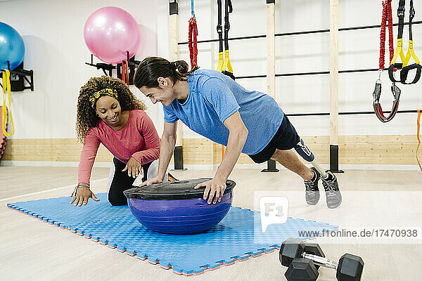 Smiling female fitness instructor assisting disabled man exercising on balance ball in gym