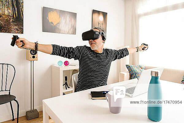 Cheerful man wearing virtual reality simulator sitting with arms outstretched at table