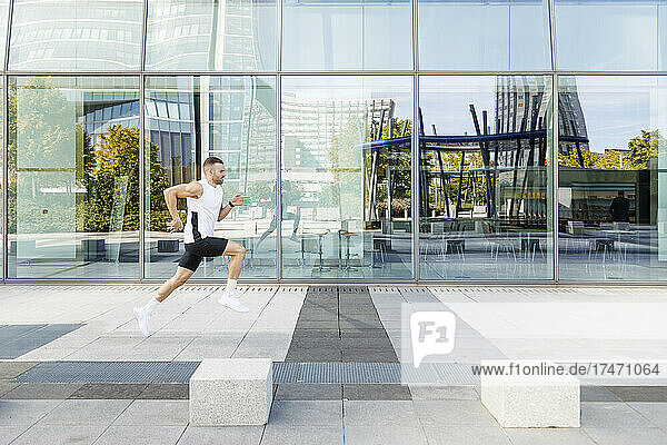 Male athlete running on footpath by glass wall